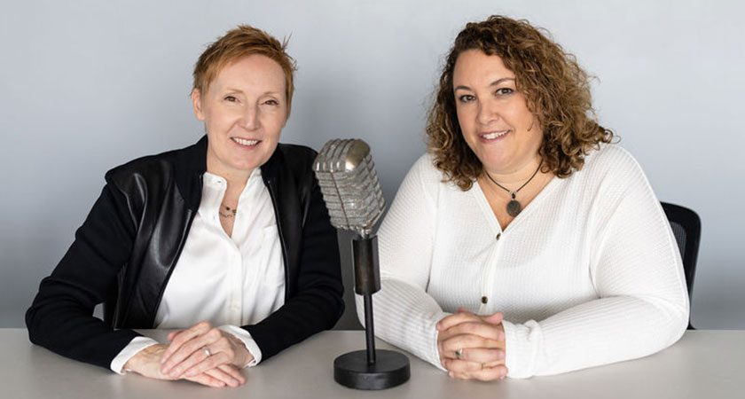 Jackie Pelland and Courtney Smock sit next to each other with their hands on a table. In between them is a classic boxy silver microphone.