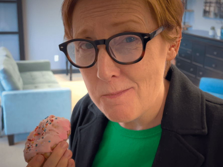 Jackie Pelland smirks at the camera as they hold a pink frosted donut in their right hand.
