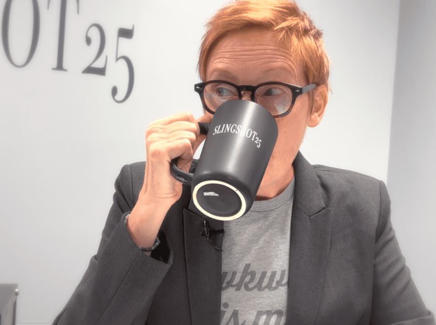 Jackie Pelland take a sip from a black mug with the "Slingshot25" logo on it. Jackie's eyes peer to the left as they sip, visible over the mug.