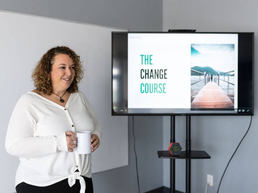 Courtney Smock stands next to a TV screen, displaying the title slide to a presentation that reads "The Change Course".