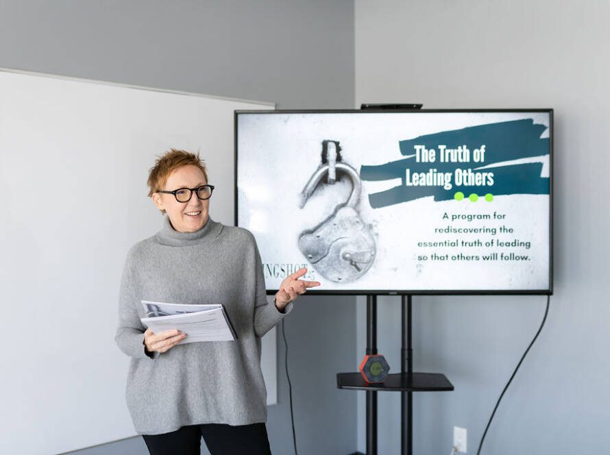 Jackie Pelland stands next to a TV screen displaying a presentation slide that reads "The Truth of Leading Others: A program for rediscovering the essential truth of leading so that others will follow".