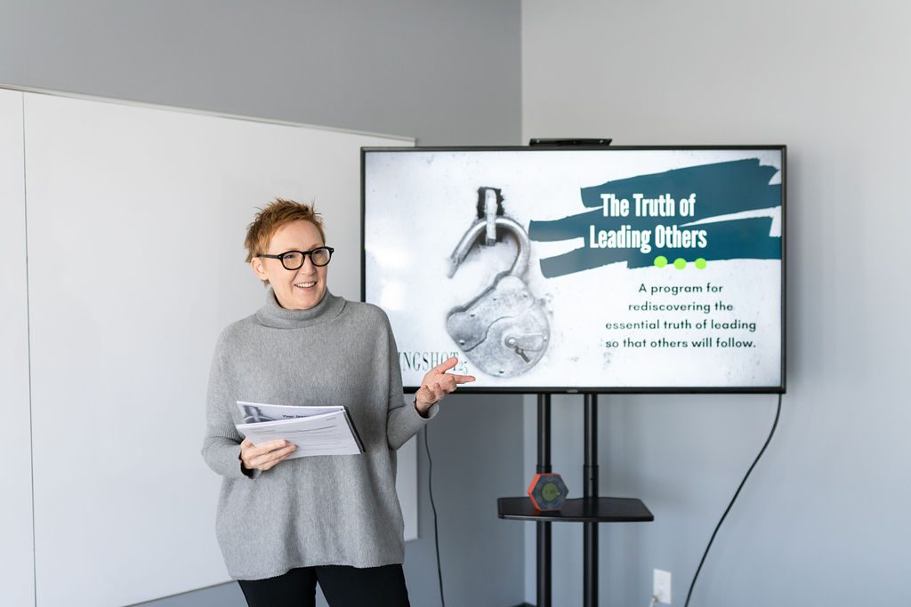 Jackie Pelland stands next to a TV screen displaying a presentation slide that reads "The Truth of Leading Others: A program for rediscovering the essential truth of leading so that others will follow".