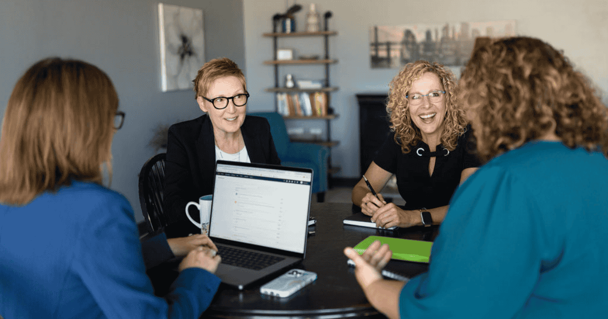 Four business women sit at a round table together with a laptop, phone, and notebooks sitting on the table