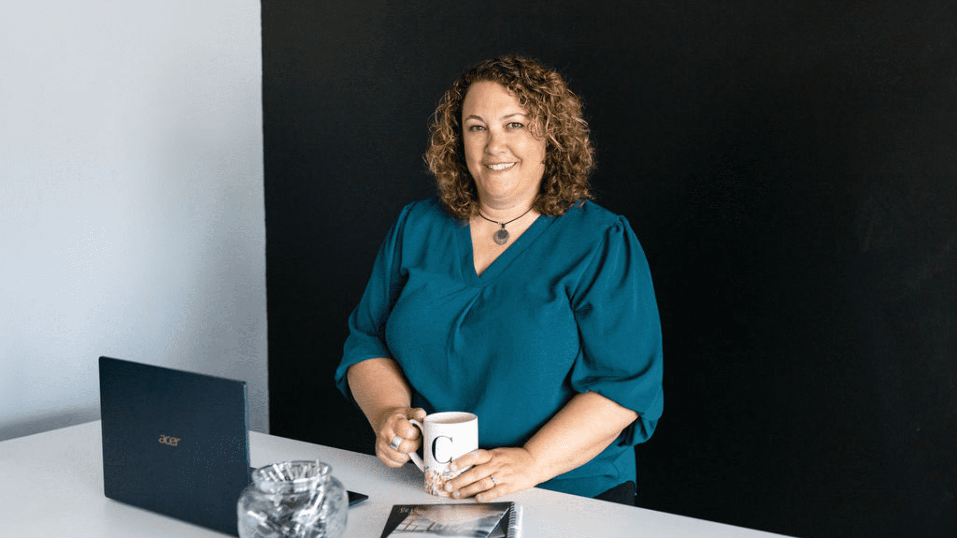 Professional female identified as Courtney Smock is seated at a desk with a coffee cup in hand, smiling with her laptop placed in front of her
