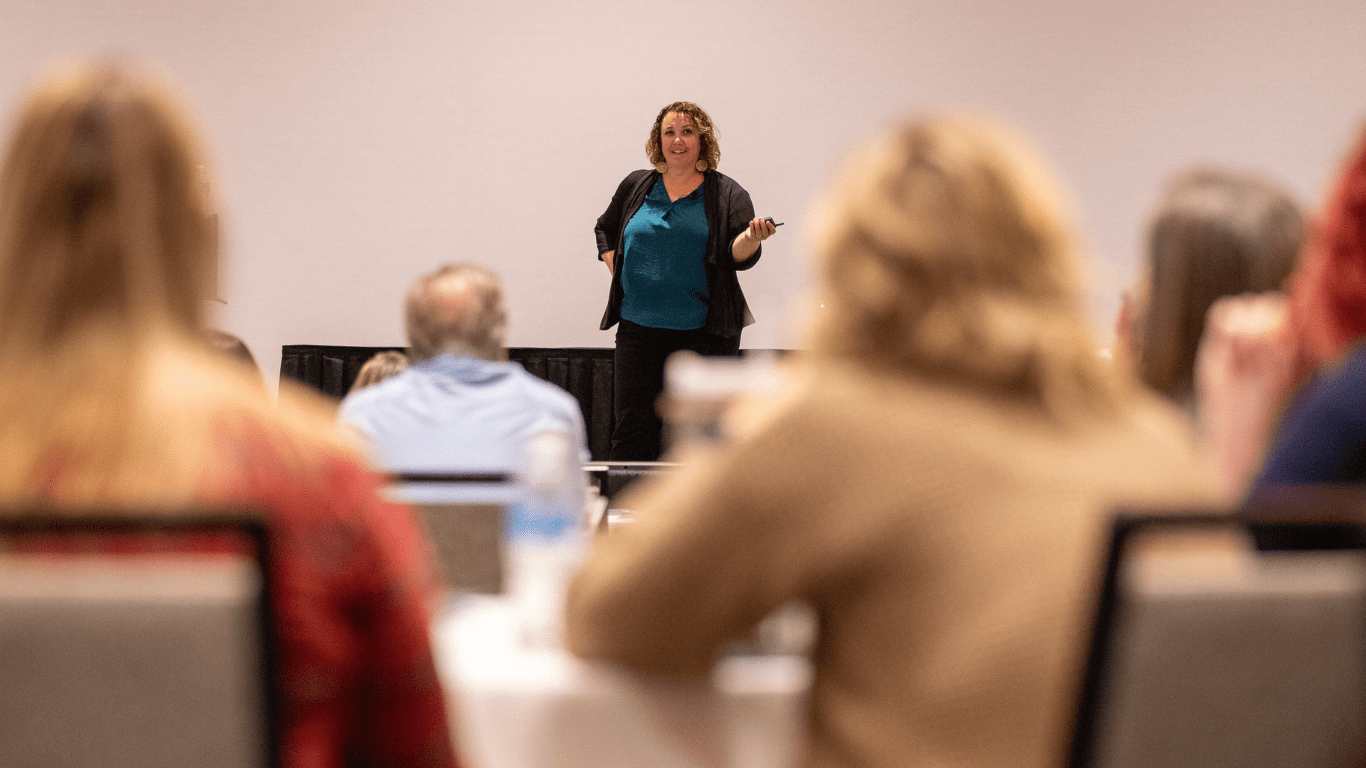 Courtney Smock - Slingshot25 Speaking Gigs Courtney Smock stands on stage in front of corporate staff meeting while guests are seated at round tables in the foreground of the photo